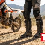 Piloto de enduro con botas de moto off-road negras junto a una moto de trail en pista de tierra